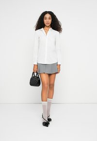 Camicia bianca a maniche lunghe, gonna grigia plissettata, borsa nera, calze al ginocchio bianche trasparenti e scarpe con dettaglio a fiocco nero. Design semplice, taglio moderno.