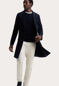 Marineblauwe wollen jas met een reverskraag, enkellangs ontwerp en knoopsluiting. Draagt over een zwarte trui en crèmekleurige broek.