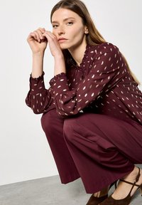 Burgundy blouse with gold leaf pattern, ruffled neckline and sleeves. Paired with matching wide-leg pants and brown heeled shoes.