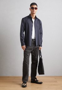 Camicia blu con bottoni sopra una t-shirt bianca, abbinata a pantaloni grigio scuro e scarpe nere con dettagli unici. Borsa nera tenuta nella mano destra.