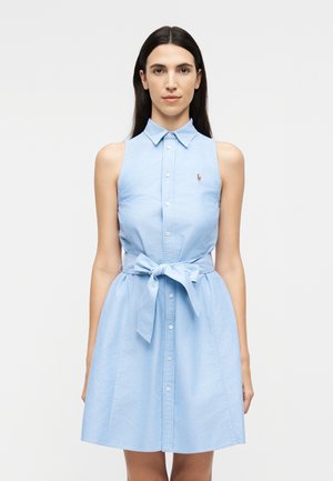 Γυναίκα με μακριά σκοτεινά μαλλιά που φοράει αμάνικο γαλάζιο φόρεμα με κουμπιά, δεμένο στη μέση με φιόγκο, στέκεται μπροστά σε απλό λευκό φόντο.