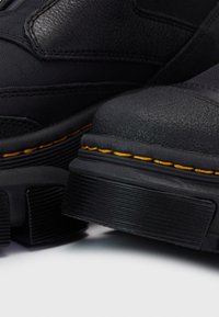 Gros plan de bottes en cuir noir avec des semelles épaisses et une couture jaune distinctive le long des bords sur fond blanc.