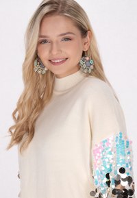 Cremfarbener Pullover mit hohem Kragen, ausgestattet mit langen Ärmeln, die mit mehrfarbigen Pailletten in Pink, Blau und metallischen Tönen verziert sind. Große blumige Ohrringe.