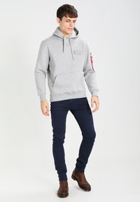 Felpa grigia con tasca frontale, cappuccio regolabile e patch del logo visibile. Abbinata a jeans blu navy e stivali alla caviglia marroni.