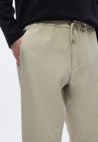 Bézs elasztikus derékpántos nadrág zsinóros rögzítéssel, oldalzsebekkel és sima anyagú textúrával. Fekete hosszú ujjú felsővel viselve.