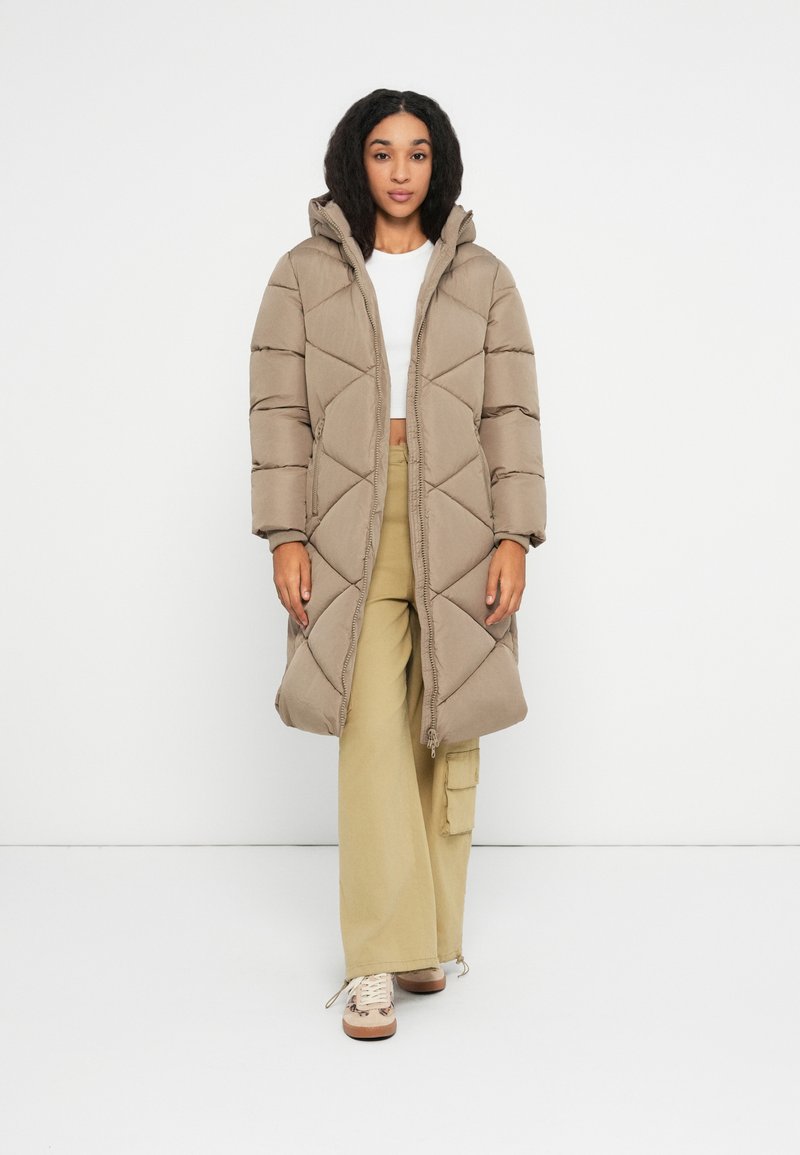 Femme portant un long manteau matelassé beige, un crop top blanc, un pantalon cargo large kaki et des baskets beiges, se tenant contre un fond blanc.