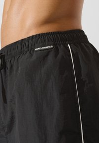 Fekete fürdőnadrág, rugalmas derékkal, fehér oldalsávval és fekete címkével, amelyen a "KARL LAGERFELD" felirat olvasható. Simább tapintás.