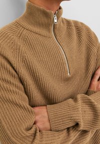 Stickad tröja i beige med hög krage och en silverdragkedja. Har ribbat mönster och långa ärmar med vida manschetter.