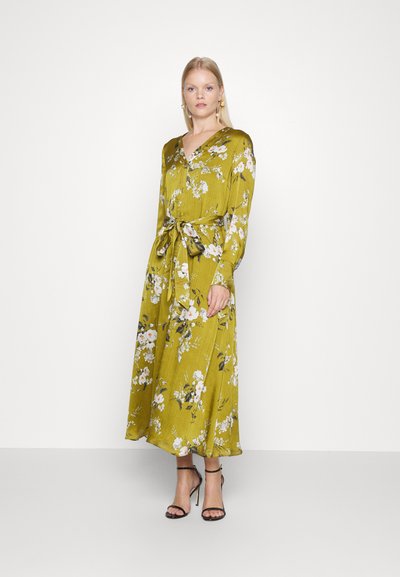 Vestuário floral de mangas longas em verde olive com um design cruzado. Apresenta um laço na cintura e uma silhueta fluida, complementada por saltos pretos.