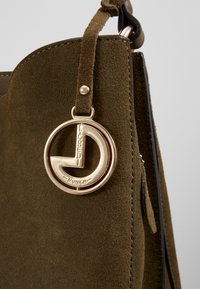 Borse in suede verde oliva con un pendente circolare in tonalità oro e una finitura testurizzata, dotata di una zip laterale.