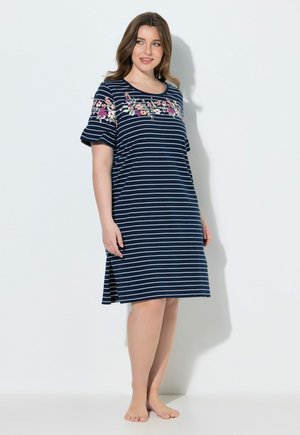 Vrouw staat blootsvoets en draagt een marineblauw-wit gestreepte jurk tot op de knie met een bloemenborduursel over de borst en mouwen.