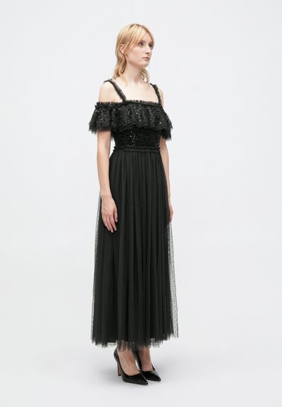 Robe noire décolletée avec des volants superposés, corsage orné et jupe en tulle fluide. Des talons hauts noirs complètent le look.