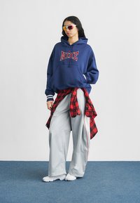 Νεαρός που φοράει μπλε-σκούρα μπλούζα Nike με κουκούλα, άσπρο φαρδύ παντελόνι, κόκκινο καρό πουκάμισο δεμένο στη μέση, άσπρα αθλητικά παπούτσια και αθλητικά γυαλιά ηλίου.