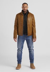 Braune Lederjacke mit hohem Kragen, kombiniert mit einer schwarzen inneren Schicht. Getragen mit hellblauen Jeans und braunen Schnürstiefeln.