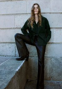 Dark green oversized shirt, high-waisted black leather pants, and ankle boots with a block heel, set against a stone wall background.