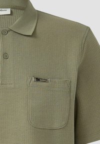 Leicht olivgrünes, strukturiertes Polohemd mit einer Brusttasche und einem kleinen Metall-Label "5.0 Elevated Casual" auf der Tasche.