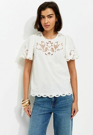 Donna che indossa una camicetta bianca ricamata a maniche corte con bordi smerlati e jeans blu, accessoriata con braccialetti dorati.