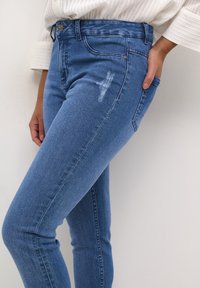 Mujer lleva unos pantalones ajustados de mezclilla azul claro con un pequeño parche desvaído en el muslo y una camiseta blanca de manga larga con canalé, con la mano en el bolsillo.