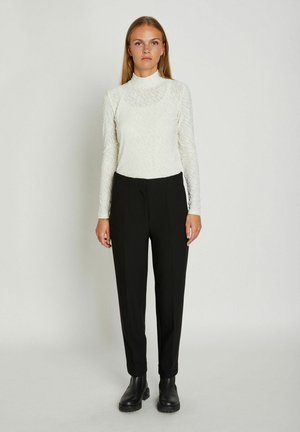 Kvinde stående iført en hvid tekstureret bluse med lange ærmer, sorte skræddersyede bukser og sorte ankelstøvler på en ensfarvet baggrund.