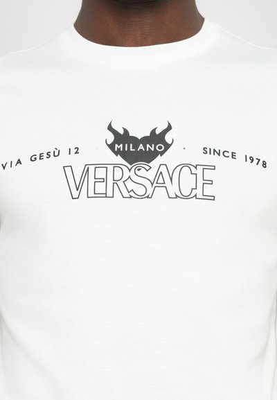 Valge puuvillane T-särk musta graafika tekstiga "VERSACE", millel on tulemotivid ja "MILANO" peal ning "SINCE 1978" all.