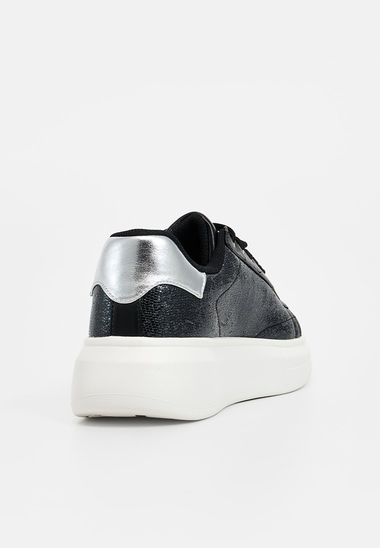 Zapatillas negras texturizadas con suela blanca, que presentan un acento metálico plateado en el talón y cordones negros, vistas desde la parte trasera.