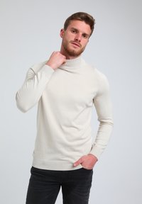 Bež turtleneck pulover iz mehkega pletenega materiala, prilegajoča se zasnova, z dolgimi rokavi ter rebrastimi manšetami in robom, v kombinaciji s temnimi kavbojkami.