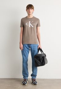 Rudas medvilninis marškinėlių su apykakle modelis su baltu Calvin Klein Jeans logotipu, derinamas su mėlynomis džinsais ir juodu sportiniu krepšiu.