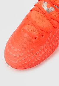 Bota de fútbol de color naranja brillante con parte superior de malla texturizada, diseño con cordones y logotipo plateado. Presenta un patrón de puntos en la suela.