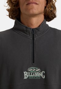 Sweat-shirt gris à col montant avec fermeture éclair sur le devant, orné du logo brodé "Billabong" en blanc et turquoise. Tissu doux et texturé.