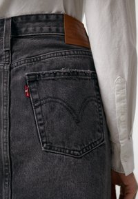 Gros plan d'une personne portant un jean en denim noir avec un patch en cuir Levi's et une chemise blanche à manches longues.