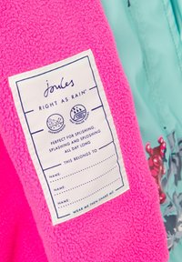 Fleecejakke i lys pink med et vævet mærke, der indeholder plejevejledninger. Mærket er hvidt med marineblå tekst, der bekræfter, at det er vandtæt.