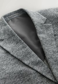 Šedý vlněný blazer s texturovaným povrchem a hladkým šedým podšívkou. Má sklápěcí límec a přiléhavý střih.