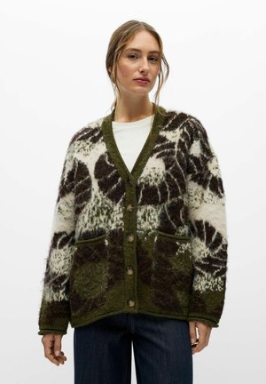 Un mannequin porte un cardigan ample à motifs olive, crème et marron sur un haut blanc et un jean foncé, devant un fond blanc uni.