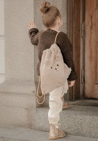 Beige plyschväska formad som en björn, med dragskoöppning upptill och ljusbruna remmar. Bärs på ett barns rygg, kombinerad med krämfärgade byxor och glittriga stövlar.