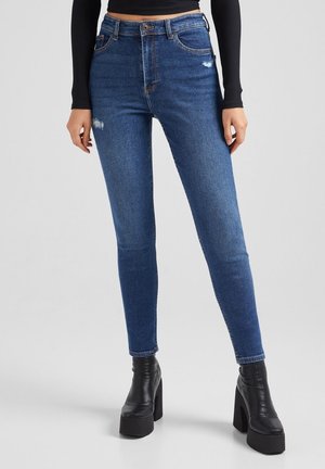 Femme portant un jean skinny bleu taille haute avec un léger effet usé et des bottines noires à talons plateforme sur un fond uni.