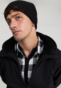 Beanie a coste nera, felpa nera con zip e colletto foderato in sherpa, indossata sopra una camicia a scacchi bianco e nero con dettagli a bottoni.