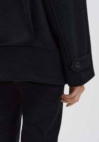 Cappotto nero dalla texture strutturata, con polsini bottonati e tasca laterale, abbinato a pantaloni neri.