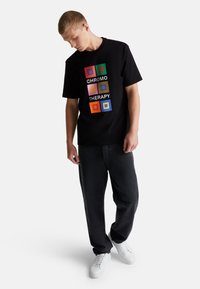 Camiseta de algodón negra con gráficos cuadrados coloridos y el texto "TERAPIA DE COLOR" en letras blancas en negrita. Mangas cortas, cuello redondo.