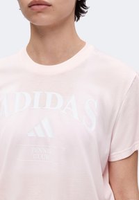 Άτομο που φοράει ένα ανοιχτό ροζ T-shirt Adidas Tennis Club με κοντά μανίκια και στρογγυλή λαιμόκοψη.
