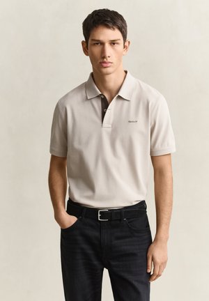 Jeune homme portant un polo beige avec le logo "GANT" et un jean noir, debout devant un fond clair uni, une main dans la poche.