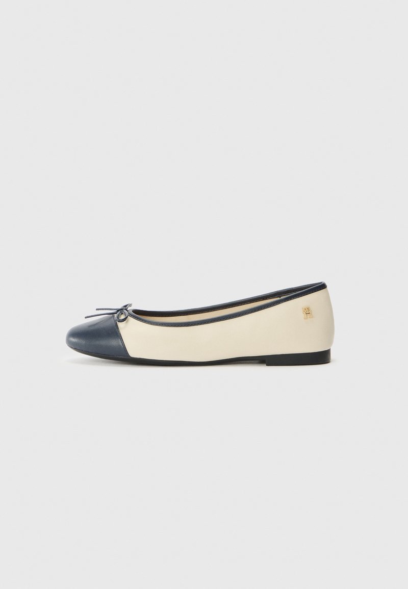 Chaussure de ballet plate beige et marine avec un petit nœud sur l’orteil marine et un logo doré près du talon sur fond blanc.