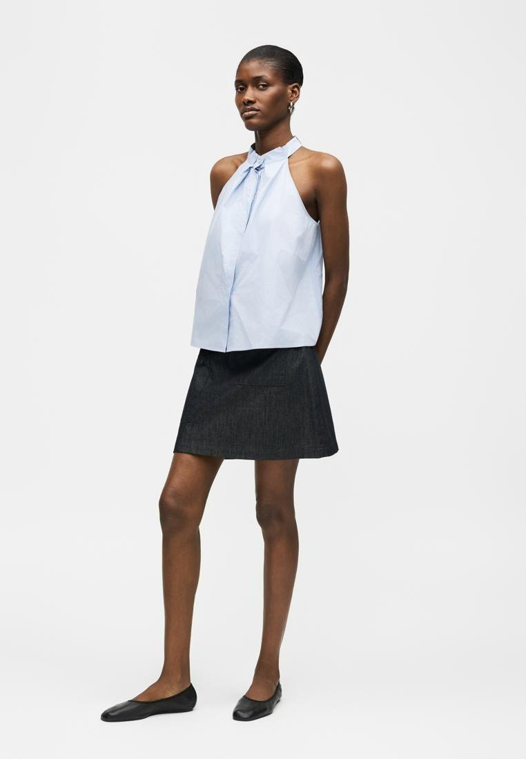 Femme portant un haut bleu clair sans manches à col licou, une jupe évasée foncée et des ballerines noires, debout devant un fond blanc uni.