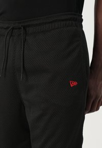 Črne mrežaste kratke hlače z elastičnim pasom, ki vključujejo rdeč logo na spodnjem delu noge ter podrobnosti s vrvico. Strukturoirana in zračna tkanina.