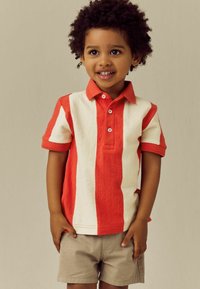 Rød og creme stribet kortærmet polo shirt med krave, parret med beige shorts. Har knapdetaljer på stolpen.