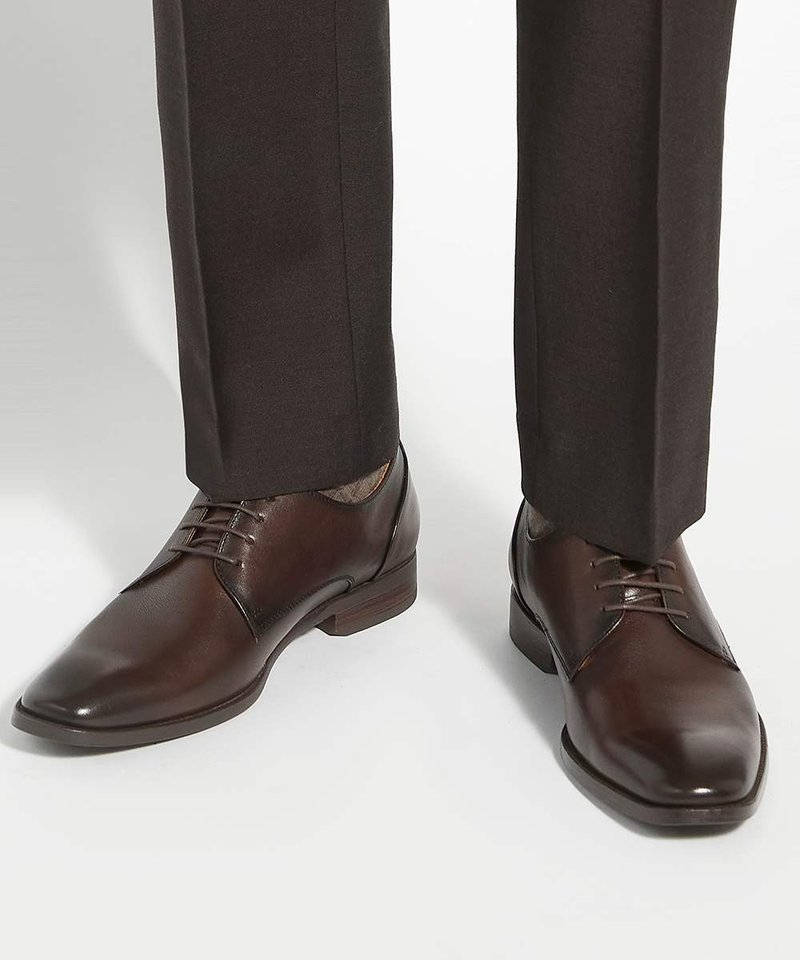 Chaussures de ville en cuir brun foncé avec lacets, portées avec un pantalon foncé sur un fond clair uni.