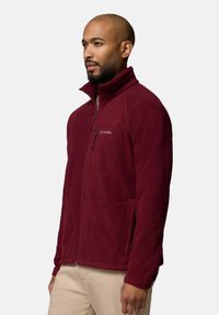 Giacca in pile bordeaux con colletto alto, chiusura con zip e tasche laterali. Texture morbida e branding minimale sul lato sinistro del petto.
