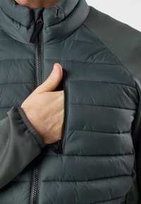 Χέρι που τοποθετεί τον αντίχειρα μέσα σε μερικώς ξεκουμπωμένη τσέπη στο στήθος ενός σκουρογκρι μπουφάν με επένδυση, μακριά μανίκια και φερμουάρ μπροστά.