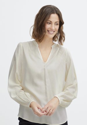 Blusa di crema chiara realizzata in un tessuto liscio e trasparente. Caratterizzata da un profondo scollo a V, maniche lunghe arricciate e una leggera texture scintillante.