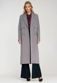 Szary płaszcz w kratę hounds-tooth zapinany na podwójne guziki, z pikowanymi klapami, bocznymi kieszeniami i luźnym krojem, noszony na bordowej bluzce z falbanami oraz ciemnych spodniach.