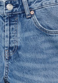 Gros plan d'un jean en denim bleu clair montrant la poche avant, la passants de ceinture, le bouton en métal et les détails de couture orange.